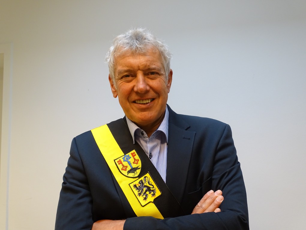 Man met grijs haar draagt een donker pak en een geel-zwarte sjerp met wapenschilden, lacht en heeft armen gekruist. (vergroot foto)