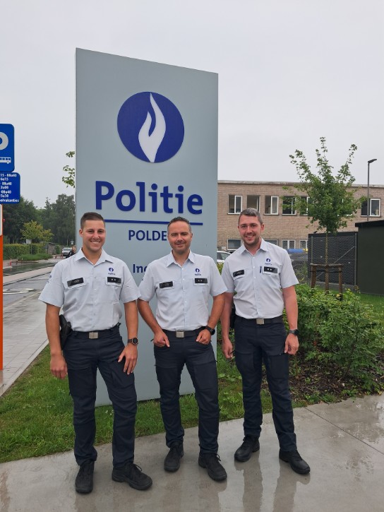 Drie politieagenten in uniform poseren lachend voor een politiebord bij een politiebureau. (vergroot foto)