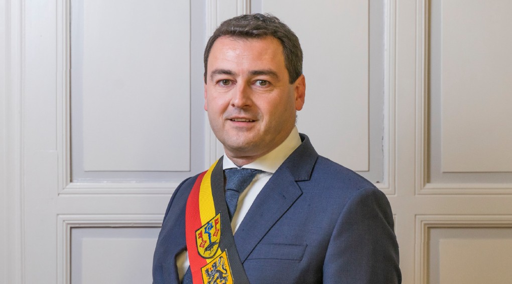 Een man in een donkerblauw pak met een witte overhemd en een blauwe stropdas, die een sjerp met het Belgische wapenschild draagt, staat voor een witte paneeldeur en kijkt in de camera. (vergroot foto)