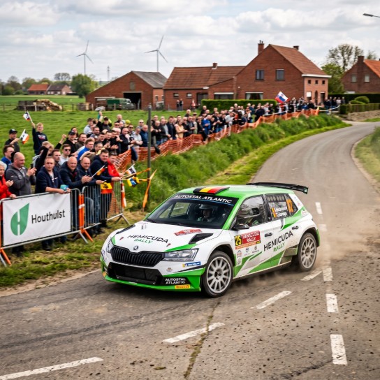 Rallyauto met groene en witte kleuren rijdt een bocht om, omringd door juichend publiek langs het platteland. (vergroot foto)