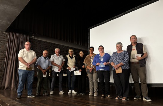 Negen mensen staan op een podium, elk met een envelop in de hand, voor een groot wit scherm. (vergroot foto)