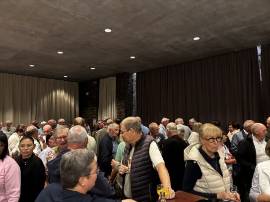 Een drukke bijeenkomst met veel mensen die met elkaar praten in een zaal met donkere gordijnen en een betonnen plafond. (vergroot foto)
